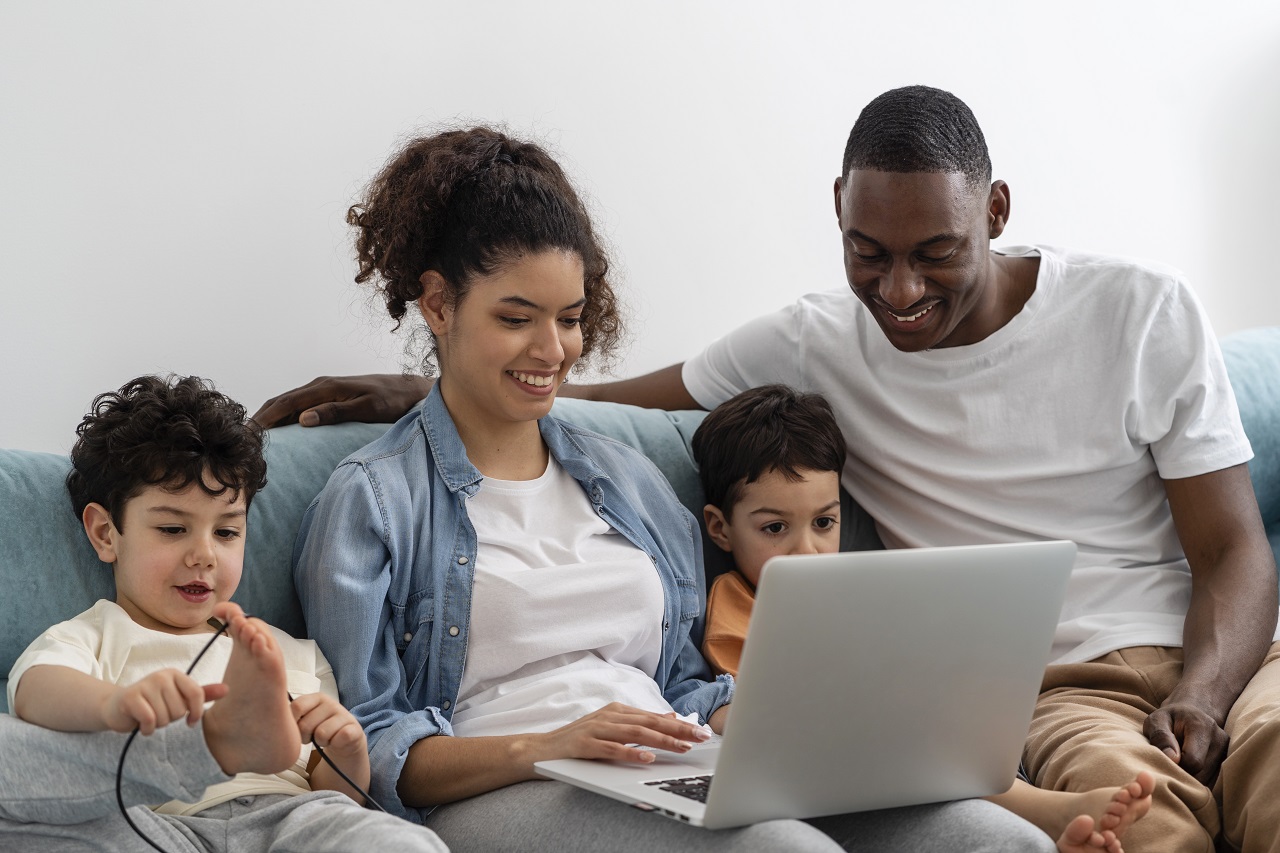 dois meninos sentados no sofá ao lado de uma mulher jovem negra e um homem jovem negro todos estão olhando um laptop e sorrindo