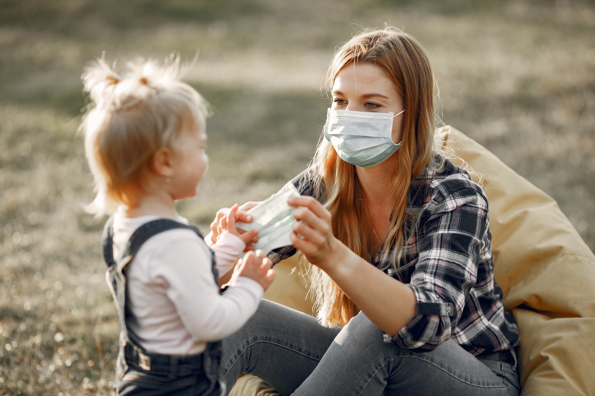 mulher sentada com uma máscara facial médica segurando outra em sua mão uma menina loira pequena está em pé na sua frente