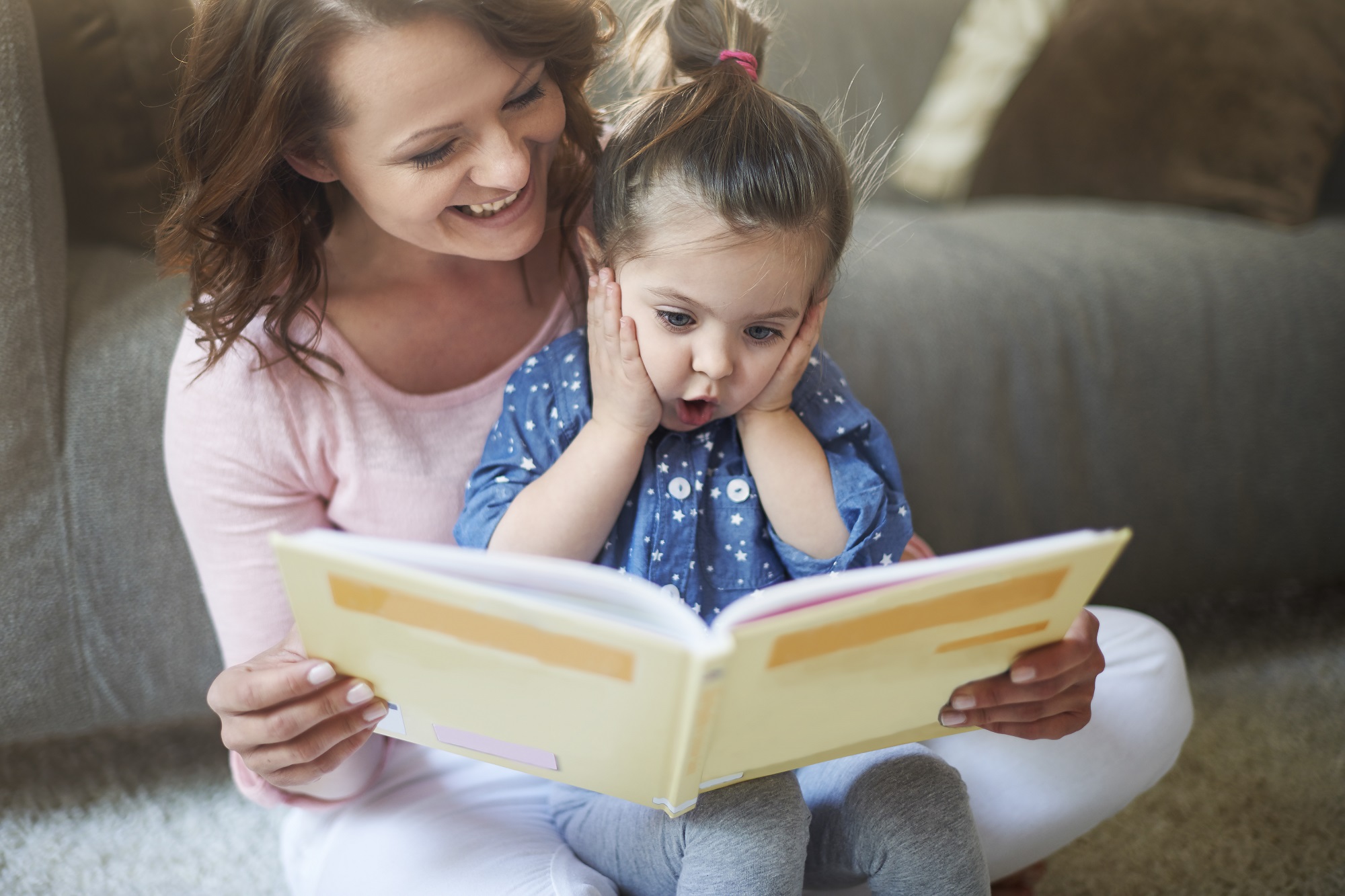 mulher sorrindo segurando um livro com uma garotinha sentada em suas pernas olhando com surpresa para o livro