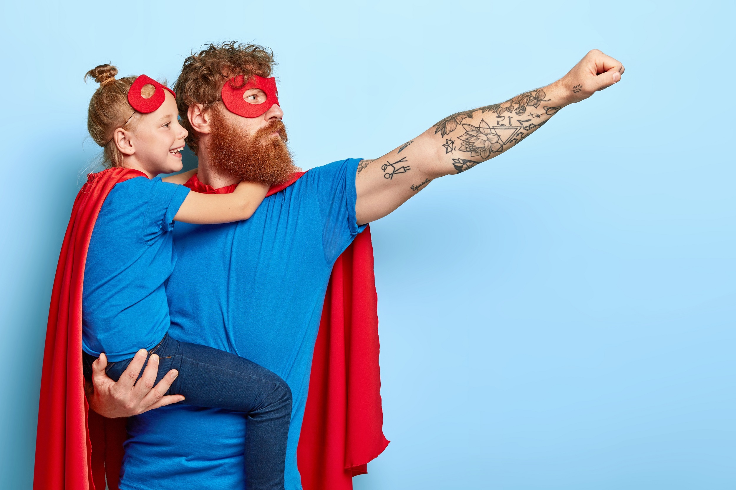 um homem ruivo grande com o braço tatuado, com uma menina loira no colo, ambos estão vestindo camisas azuis, máscara e capas vermelhas como heróis