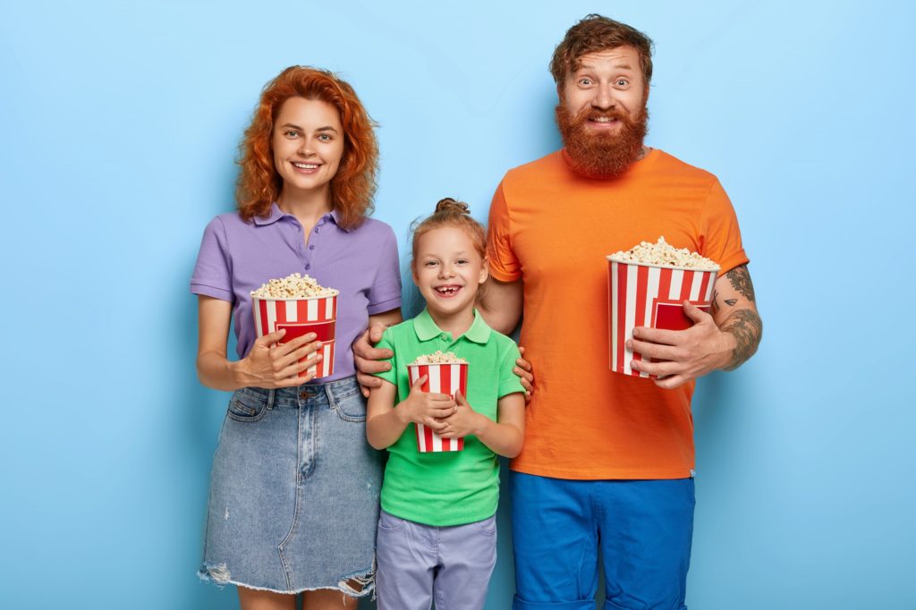 mulher, menina e homem todos ruivos com baldes de pipoca nas mãos sorrindo em um fundo azul