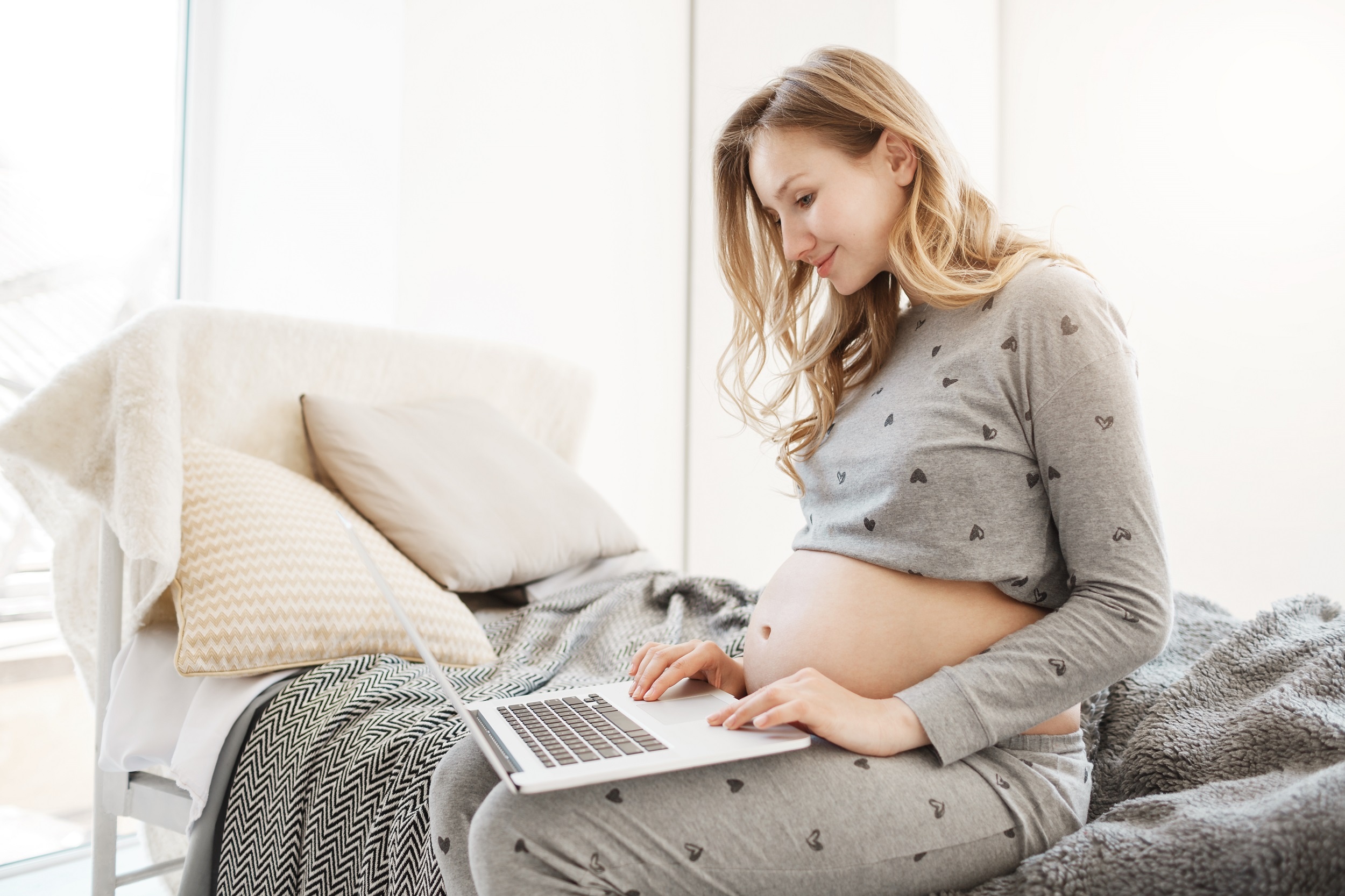 mulher jovem loira grávida sentada na cama com um notebook apoiado nas pernas