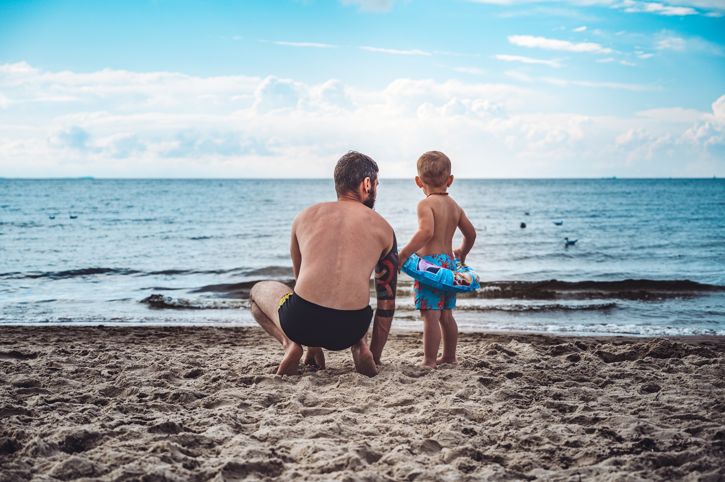 homem e um menino de costas em uma praia, o homem está agachado na areia e o menino loiro ao lado