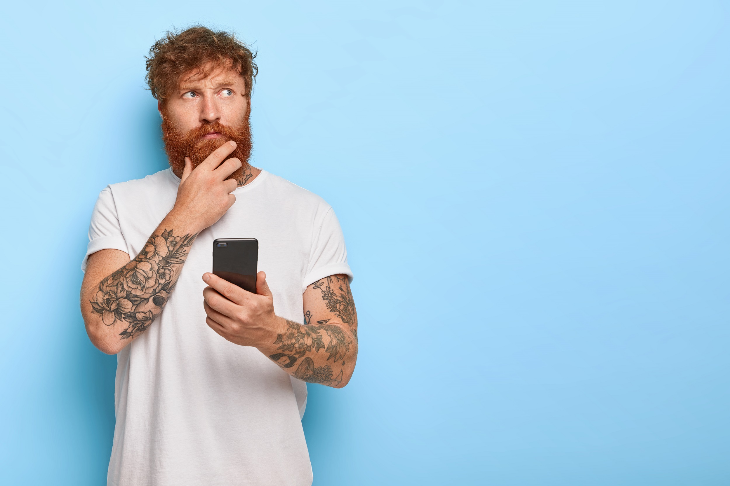 homem ruivo de camisa branca e braços tatuados segurando um celular na mão com expressão pensativa