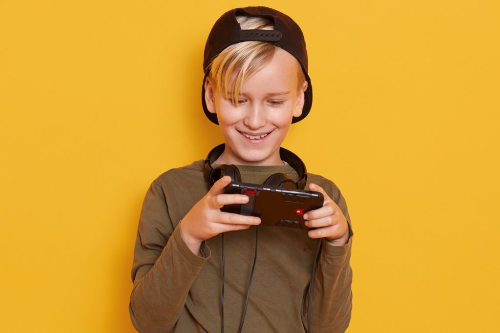 garoto loiro em fundo amarelo sorrindo com fone de ouvido no pescoço olhando para a tela de um celular