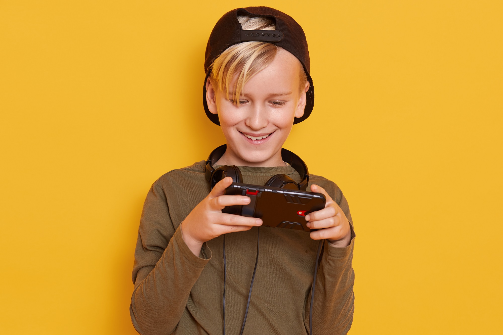 garoto loiro em fundo amarelo sorrindo com fone de ouvido no pescoço olhando para a tela de um celular