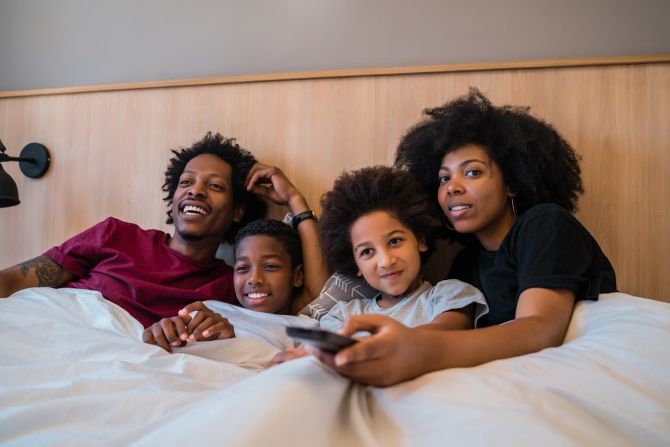 família afro-americana, um homem, um menino, uma menina e uma mulher deitados em uma cama, a mulher está com um controle remoto na mão