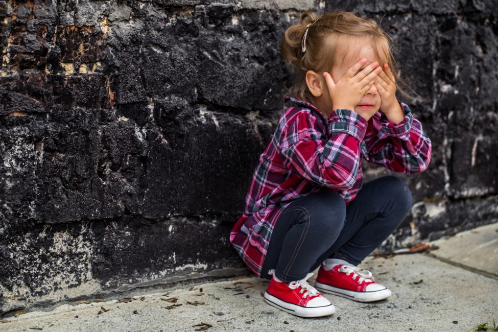 menina loira agachado no chão encostada na parede tampando os olhos com as mãos