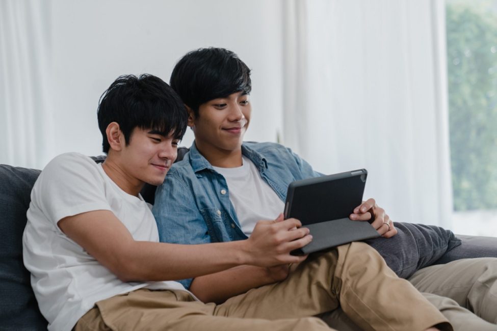 duas pessoas do gênero masculino sentados em um sofá assistindo Doramas um tablet