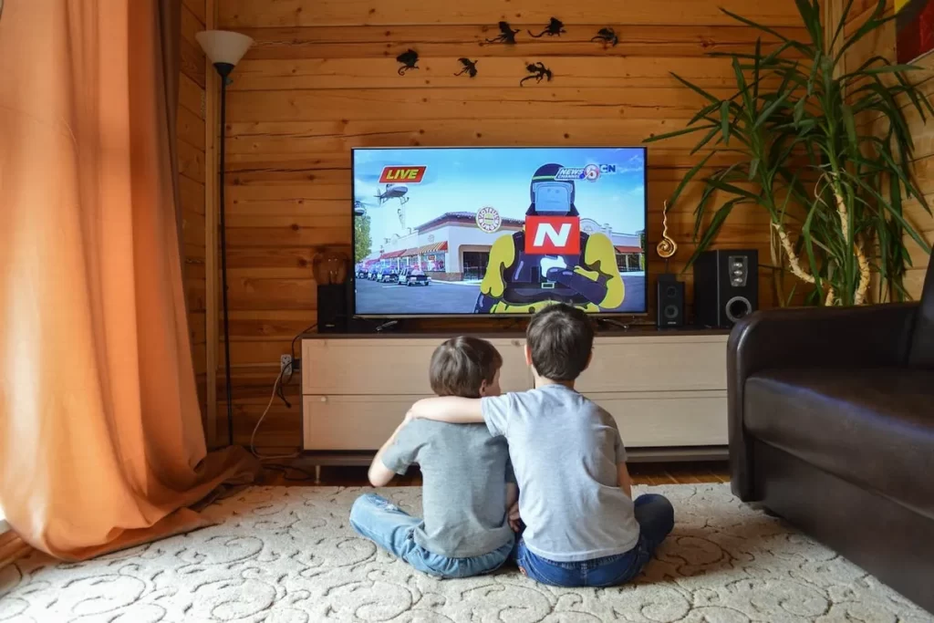 Dois meninos, um abraçando o outro, sentados no chão de uma sala aconchegante com paredes de madeira, assistindo a um desenho animado em uma TV grande.