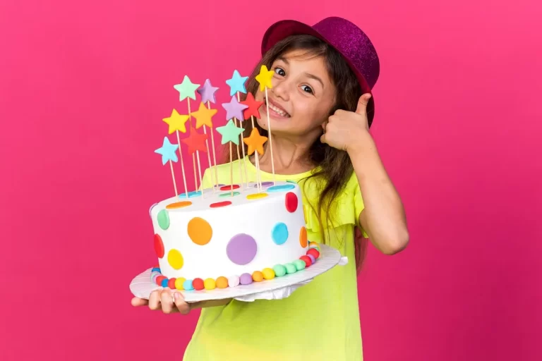 Garota feliz com chapéu roxo brilhante segurando bolo de aniversário decorado com estrelas e bolinhas coloridas, sorrindo e fazendo sinal de positivo