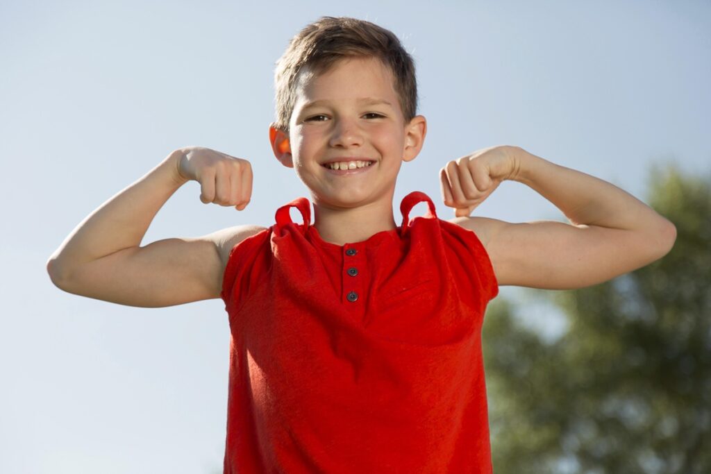 Menino sorridente usando camiseta vermelha mostra os braços fortes enquanto posa flexionando os músculos ao ar livre em um dia ensolarado.