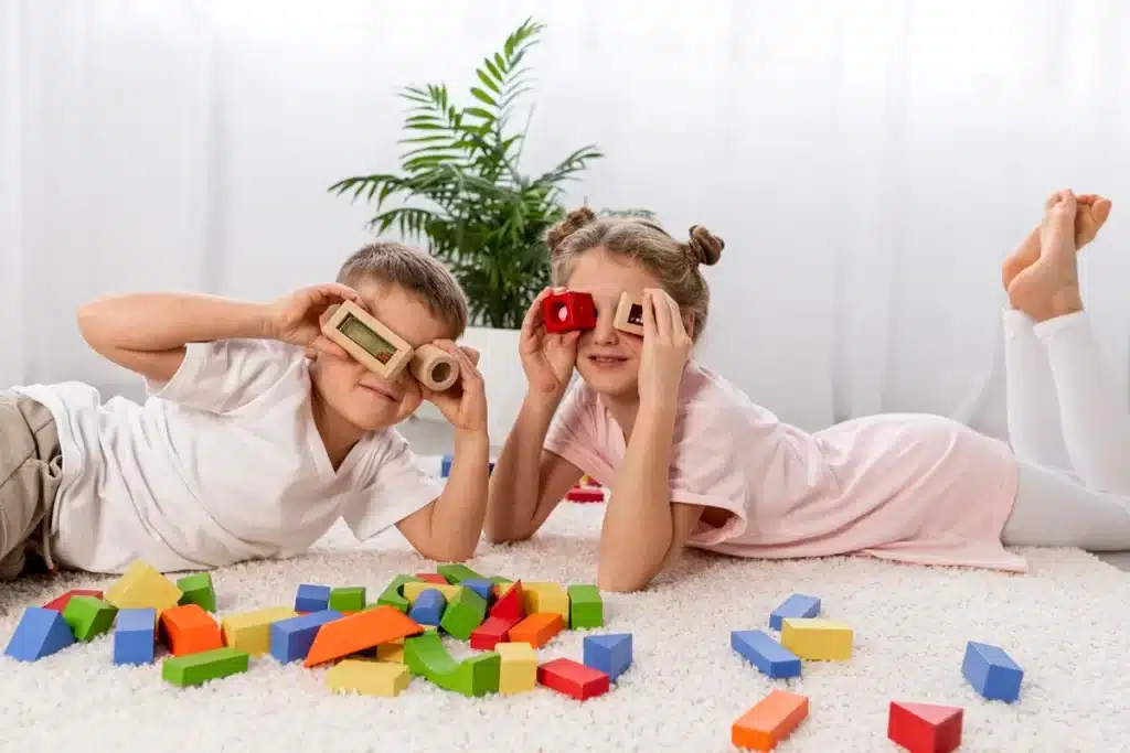 Duas crianças, um menino e uma menina, brincando com blocos de construção coloridos no chão. O menino usa um bloco de madeira como binóculo, e a menina faz o mesmo com um bloco vermelho. Ao fundo, plantas decorativas e uma sala iluminada.