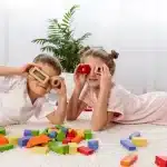Duas crianças, um menino e uma menina, brincando com blocos de construção coloridos no chão. O menino usa um bloco de madeira como binóculo, e a menina faz o mesmo com um bloco vermelho. Ao fundo, plantas decorativas e uma sala iluminada.