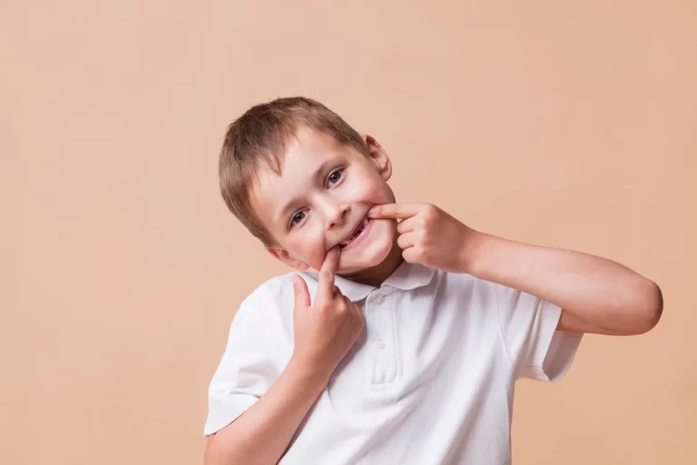 Menino fazendo careta ao puxar o canto da boca com os dedos para mostrar os dentes, vestindo camisa polo branca em fundo bege