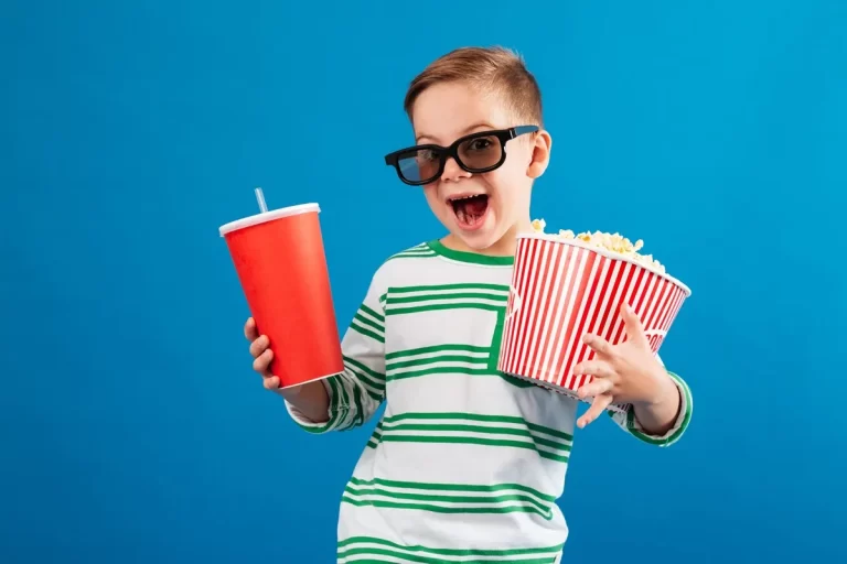 Menino sorridente com óculos 3D segurando balde de pipoca e copo de refrigerante em fundo azul, pronto para assistir a um filme