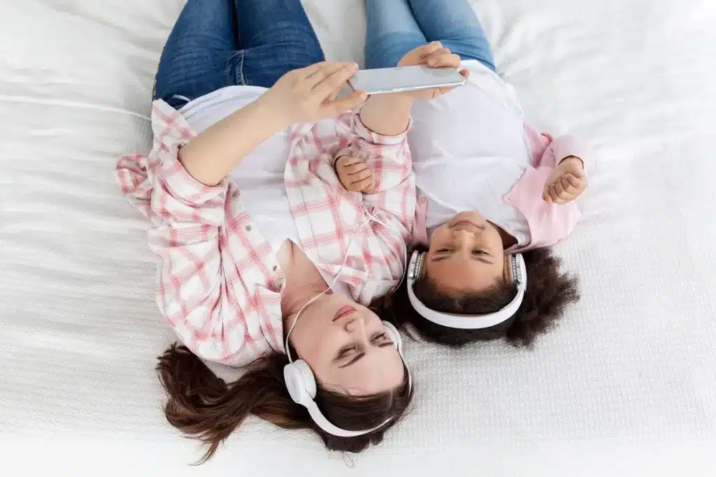 Mãe e filha deitadas juntas na cama usando fones de ouvido e olhando para celular, ouvindo música ou história para dormir