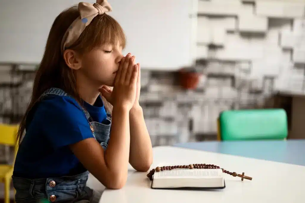 Menina em oração com as mãos juntas sobre um livro e terço.