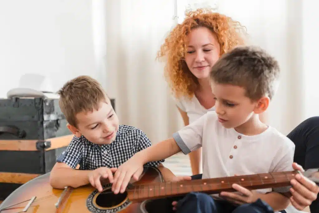 Uma mulher sorridente com cabelos cacheados ruivos (terapeuta ou mãe) observa dois meninos pequenos tocando juntos as cordas de um violão, representando o aprendizado e a musicoterapia em um ambiente claro.