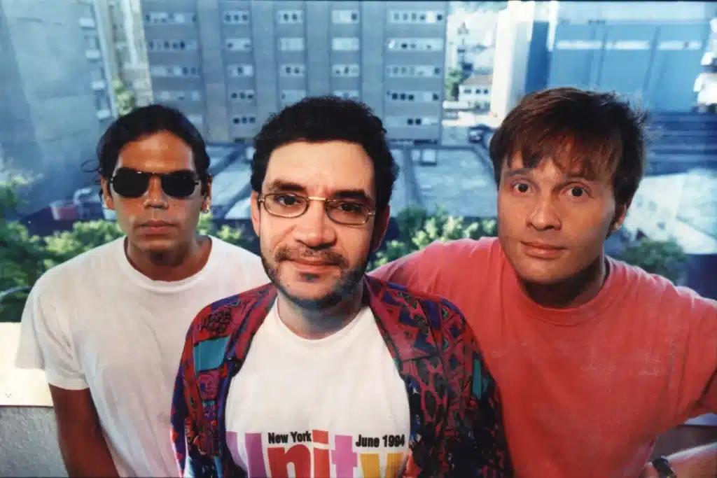 Retrato em close-up da banda Legião Urbana. Da esquerda para a direita: Marcelo Bonfá de óculos escuros e camiseta branca, Renato Russo no centro com óculos de aro fino e camisa estampada sobre camiseta branca, e Dado Villa-Lobos de camiseta vermelha.