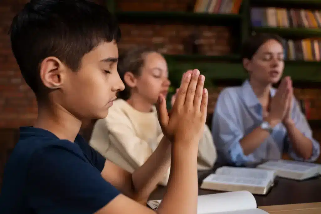 Close-up de um menino orando (mãos postas e olhos fechados) em uma mesa, com outras crianças e uma adulta em segundo plano, também em posição de oração ou meditação. Imagem representa fé e espiritualidade infantil.