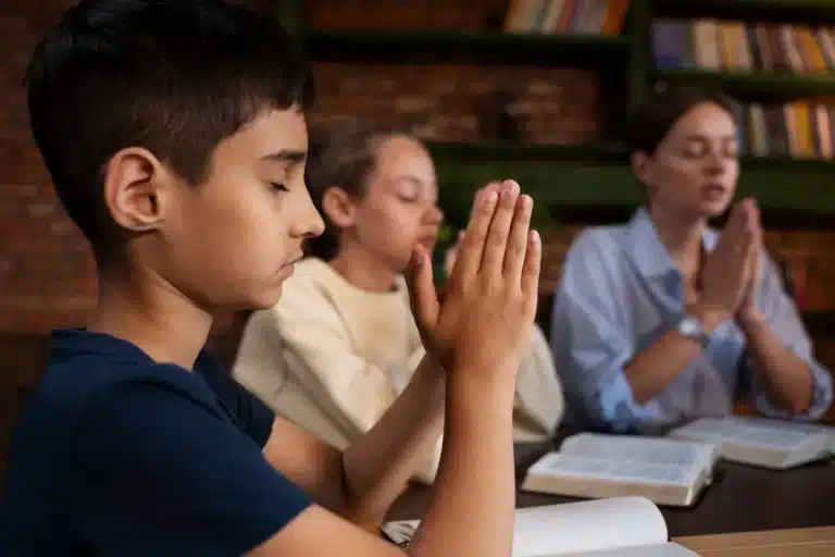 Close-up de um menino orando (mãos postas e olhos fechados) em uma mesa, com outras crianças e uma adulta em segundo plano, também em posição de oração ou meditação. Imagem representa fé e espiritualidade infantil.