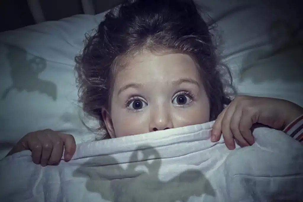 Menina assustada na cama, coberta até o rosto, com expressão de medo de pesadelos.