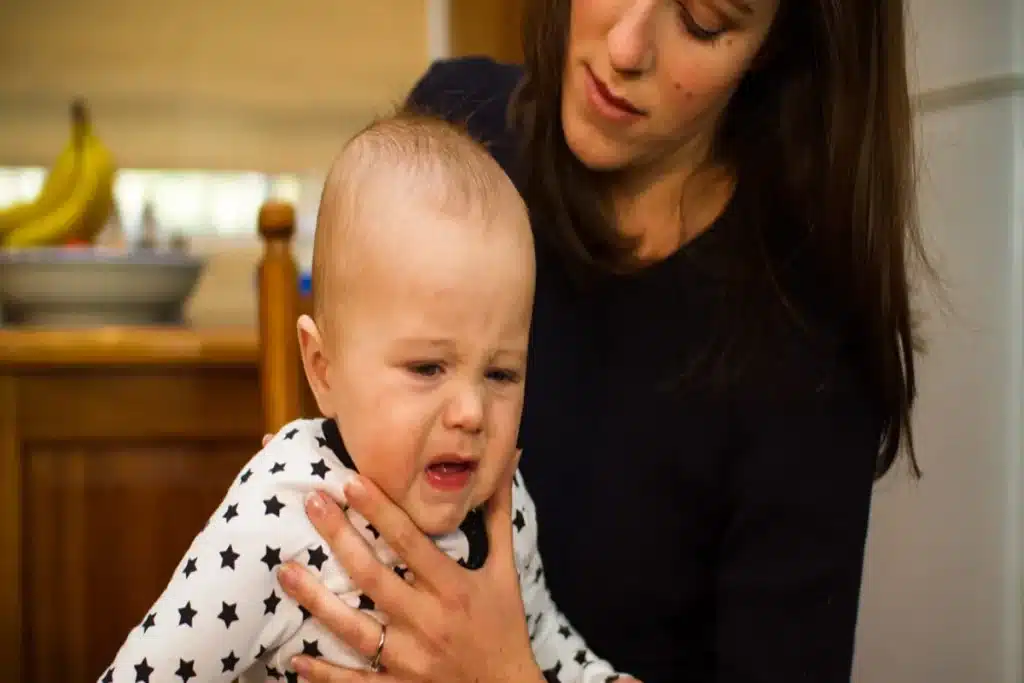 Uma mulher segura um bebê com o rosto contorcido, chorando intensamente, com suas mãos posicionadas perto do queixo da criança, em uma cena que simboliza o cuidado em um momento de aflição.