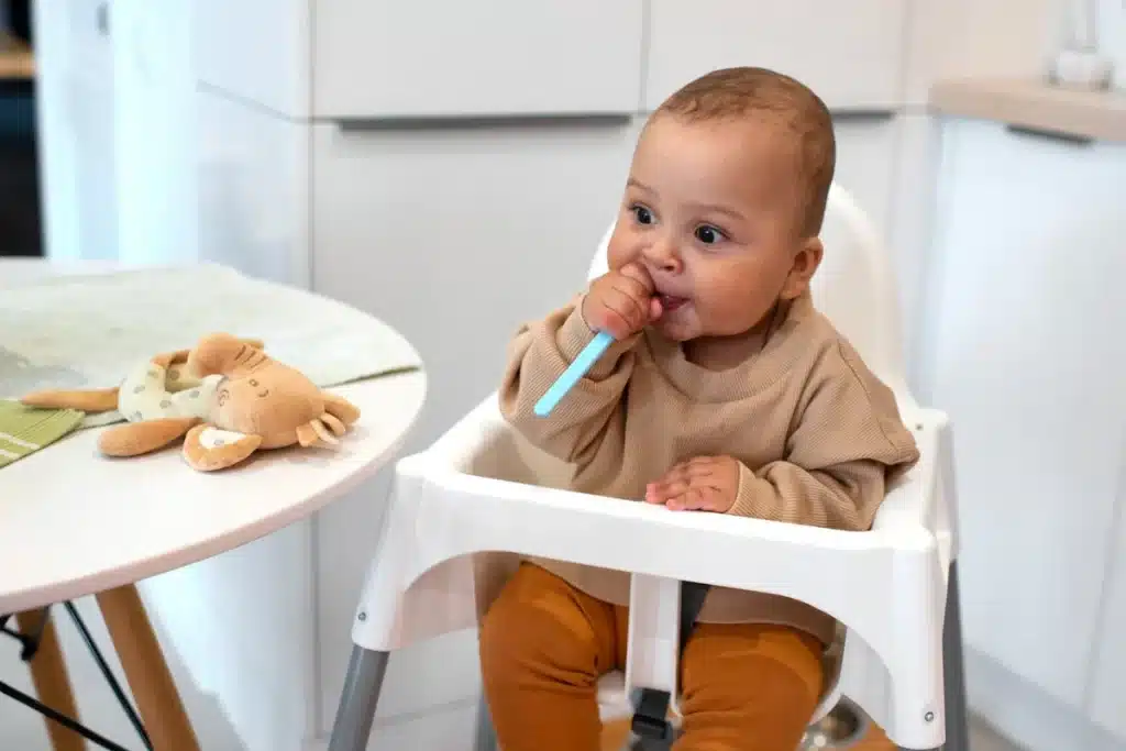Bebê sorridente sentado em uma cadeira de alimentação, mordendo uma colher de silicone azul com um ursinho de pelúcia na mesa ao lado.