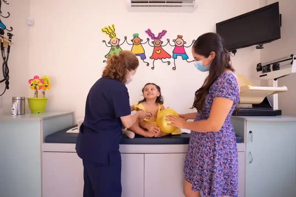Pediatra (vestindo máscara) examinando uma criança pequena e sorridente com a presença da mãe (vestindo máscara) em um consultório pediátrico decorado com desenhos infantis. A criança segura um balão amarelo.