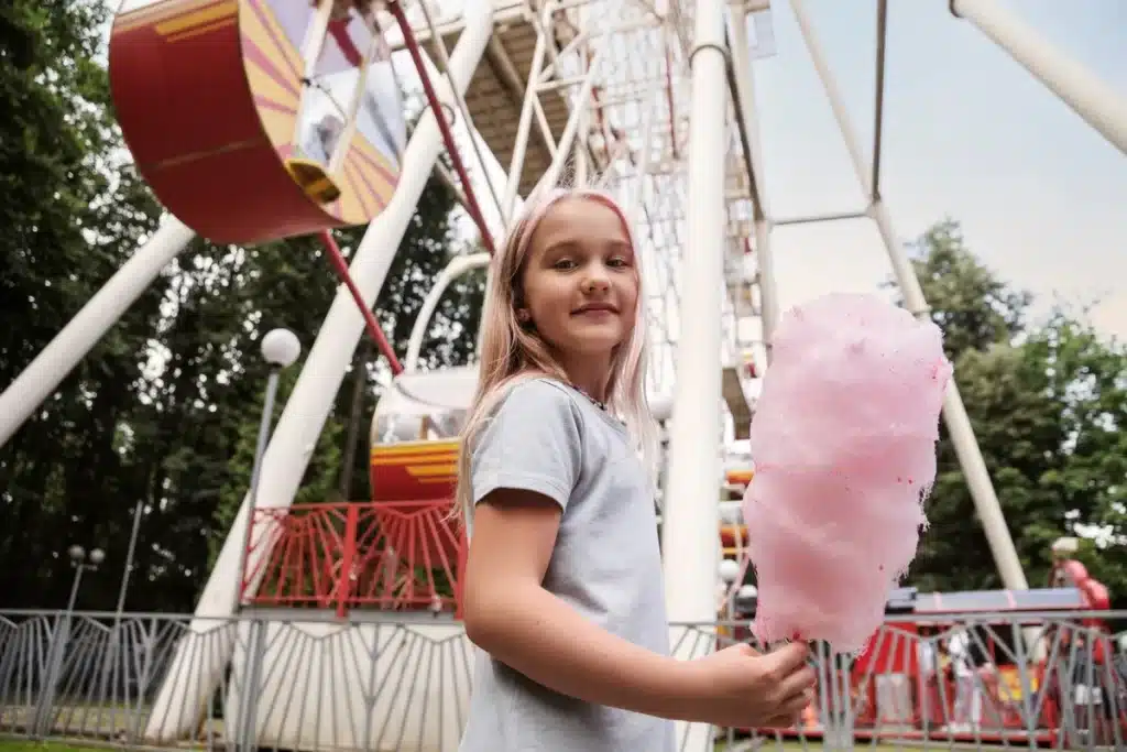Retrato de uma menina loira com pontas rosa, vestindo uma camiseta cinza, segurando um grande algodão-doce rosa e sorrindo. Ao fundo, parcialmente visível, está uma roda-gigante vermelha e branca em um parque de diversões.