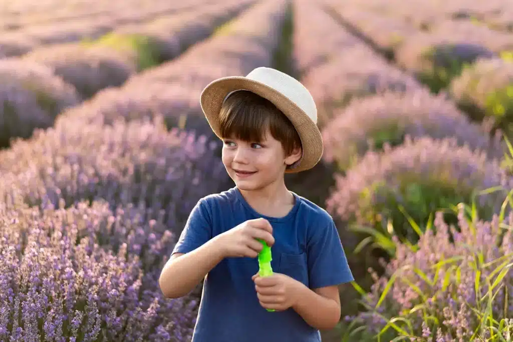 Menino sorridente com chapéu de palha brinca com um frasco de bolhas de sabão em meio a um campo florido de lavanda ao pôr do sol.