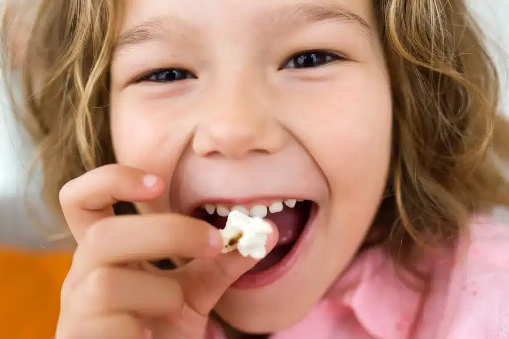 Criança feliz segurando um pedaço de pipoca e sorrindo, mostrando um dente de leite caindo, simbolizando infância e crescimento.