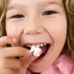 Criança feliz segurando um pedaço de pipoca e sorrindo, mostrando um dente de leite caindo, simbolizando infância e crescimento.