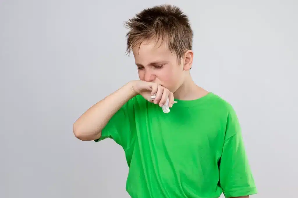 Menino com camiseta verde tossindo e cobrindo a boca com o braço, segurando um lenço, aparentando sintomas de alergia ou problemas respiratórios.