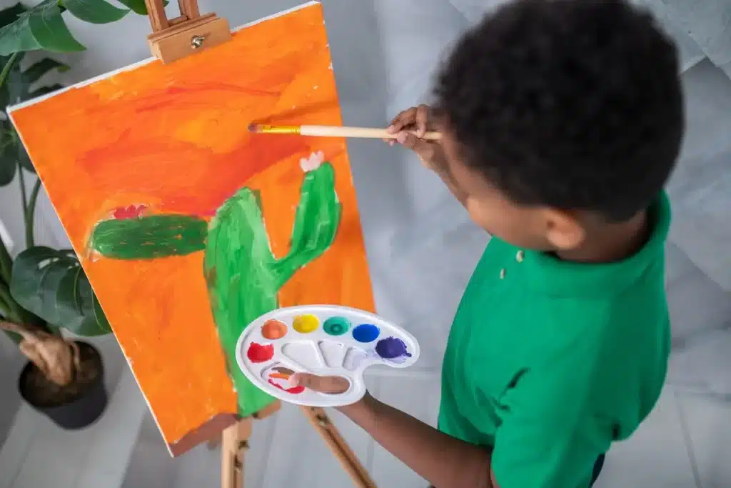 Menino segurando paleta de tintas e pincel enquanto pinta um quadro colorido com desenho de cacto.