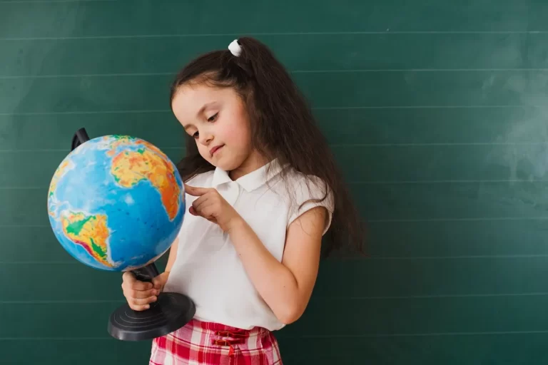 Menina com uniforme escolar apontando para um globo terrestre em frente a um quadro verde