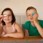 Menina e menino sorrindo enquanto brincam com peças de quebra-cabeça, cada um segurando uma peça azul sobre um dos olhos, sentados à mesa em ambiente doméstico.