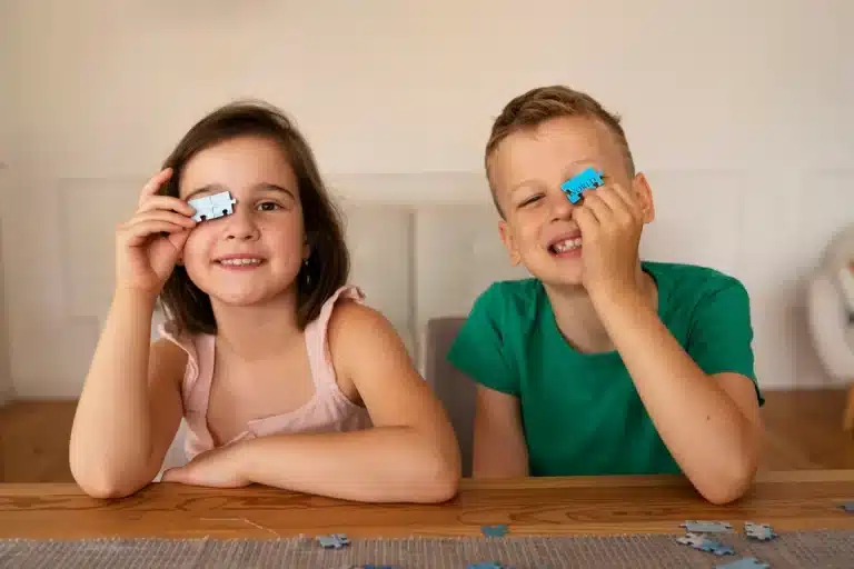Menina e menino sorrindo enquanto brincam com peças de quebra-cabeça, cada um segurando uma peça azul sobre um dos olhos, sentados à mesa em ambiente doméstico.