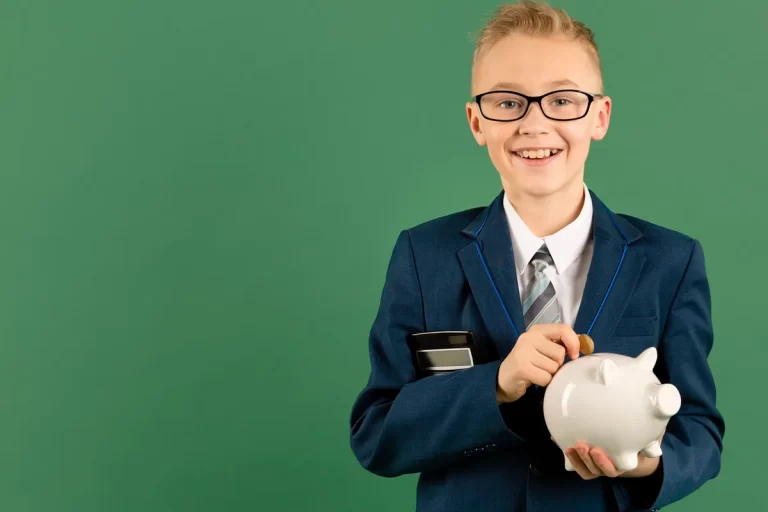 Criança com óculos e terno segurando um cofrinho branco e uma calculadora, sorrindo enquanto coloca uma moeda.