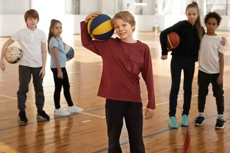 Grupo de crianças com bolas esportivas em uma quadra de madeira durante aula de educação física escolar.