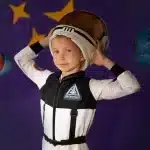 Menino com fantasia de astronauta segurando capacete espacial, com fundo roxo decorado com estrelas e planetas de papel colorido