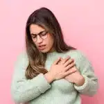 Mulher com óculos e suéter verde-claro segurando o peito com as duas mãos e expressão de dor ou desconforto, em frente a fundo rosa.