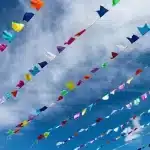 Fitas de bandeirinhas coloridas típicas de festa junina penduradas no céu azul com nuvens brancas ao fundo.