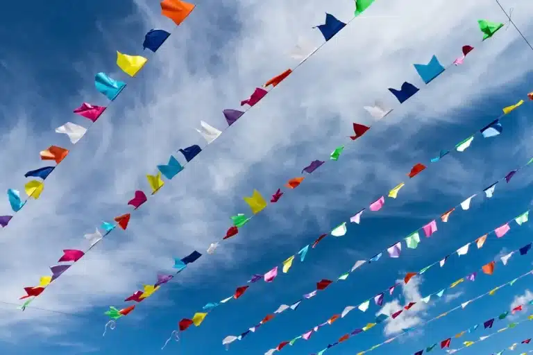 Fitas de bandeirinhas coloridas típicas de festa junina penduradas no céu azul com nuvens brancas ao fundo.