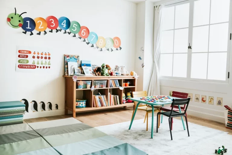 Ambiente de sala de aula para educação infantil com tapetes coloridos, estante com livros e brinquedos, mesa infantil com cadeiras coloridas e materiais educativos em cima.