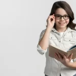 Garota com óculos de grau segurando um livro aberto e sorrindo, vestida com camisa clara e fundo branco.