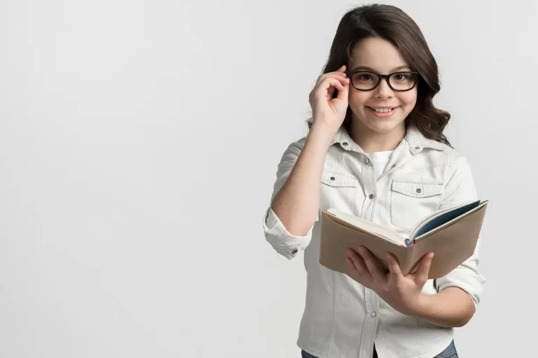 Garota com óculos de grau segurando um livro aberto e sorrindo, vestida com camisa clara e fundo branco.
