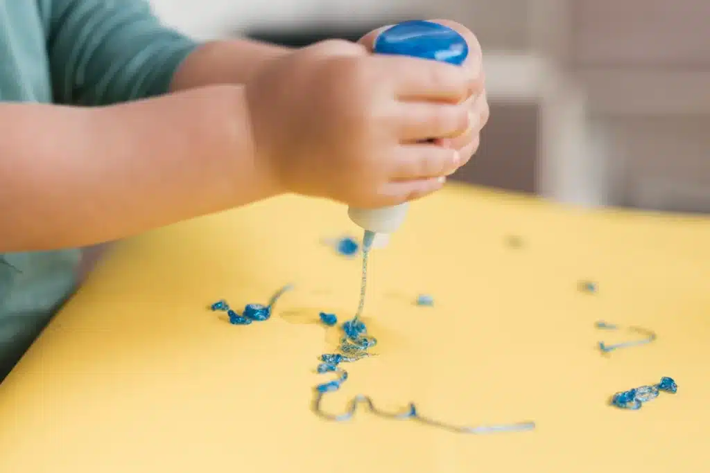 Criança apertando um tubo de tinta azul, criando linhas e formas sobre uma superfície amarela