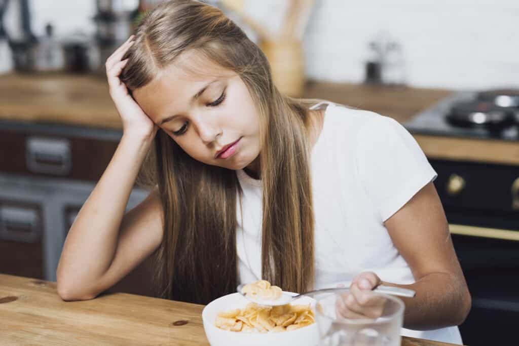Menina de cabelo longo e castanho, com uma expressão triste enquanto come cereais com leite, parecendo desinteressada e cansada. Ela está com a mão na cabeça, como se estivesse distraída ou sem vontade de comer.